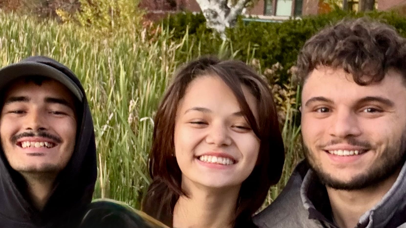Three siblings smiling brightly in front of tall reeds at Bates College, enjoying a peaceful outdoor moment