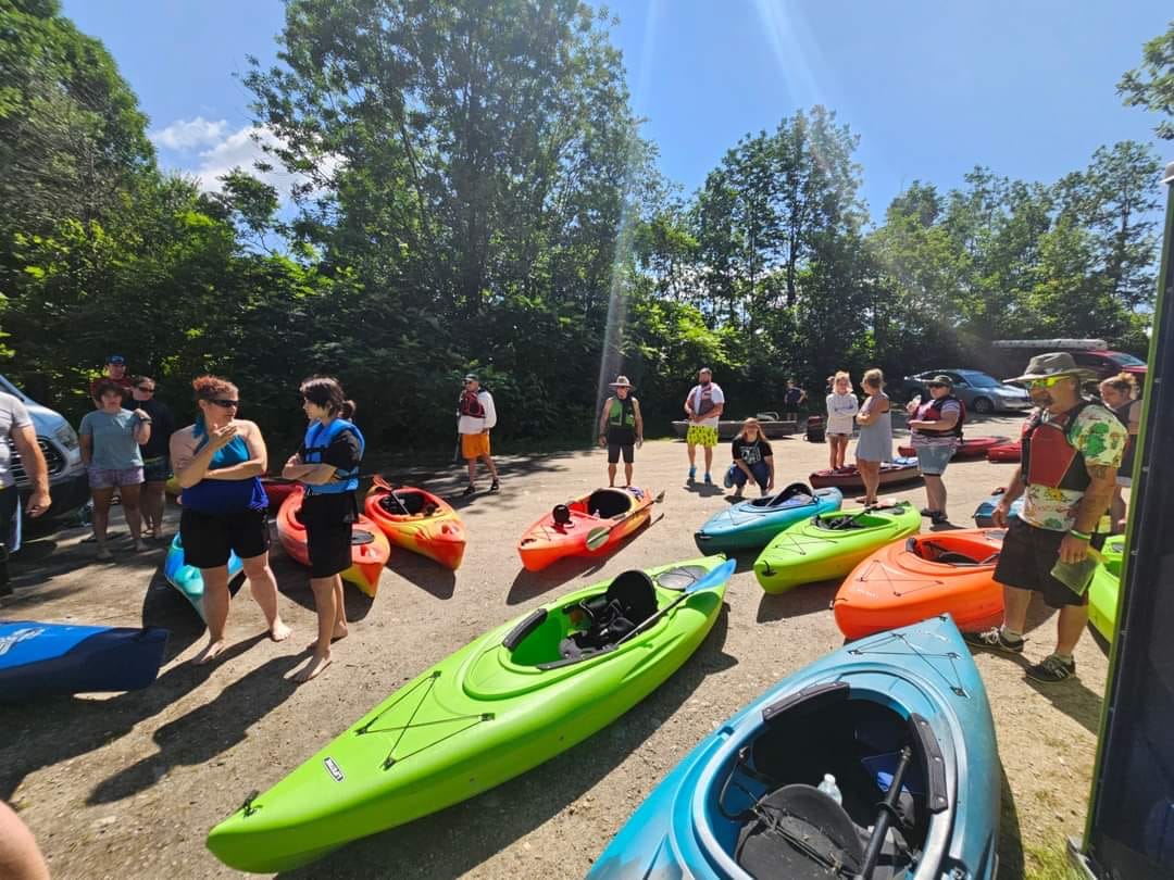 Group gathered with colorful kayaks on the Androscoggin River in Bethel, Maine, before a paddle with Western Maine Recovery.