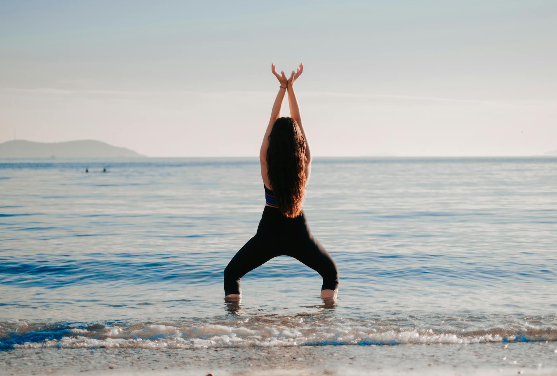 Woman practicing yoga by the ocean, standing in a grounding pose with her arms raised, embracing mindfulness and nature's serenity.