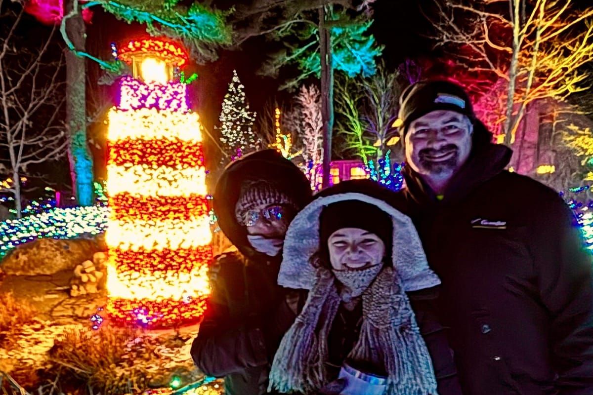 Family enjoying the holiday lights at Gardens Aglow, bundled up and smiling in front of a glowing lighthouse display.