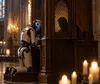 Ein humanoider Roboter kniet in einer alten Kirche vor einem hölzernen Beichtstuhl. Hinter dem Gitter ist schemenhaft ein Mensch zu erkennen.