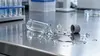 KI-generiertes Bild: Ein zerbrochenes Reagenzglas mit verschütteter Flüssigkeit auf einer Edelstahl-Arbeitsfläche in einem Labor.