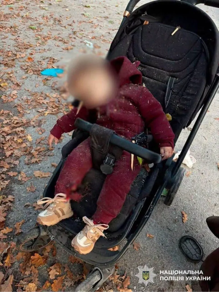 Ein getötetes Kleinkind im weinroten Overall und mit hellen Turnschuhen sitzt angeschnallt in einem schwarzen Kinderwagen auf einem Weg voller Herbstlaub. Das Gesicht ist unkenntlich gemacht.