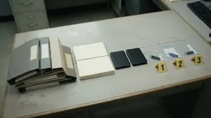 Ein grauer Bürotisch von schräg oben. Darauf liegen ordentlich aufgereiht ein Stapel grauer und beiger Aktenordner, zwei Stapel Papier, zwei schwarze externe Festplatten und drei USB-Sticks i