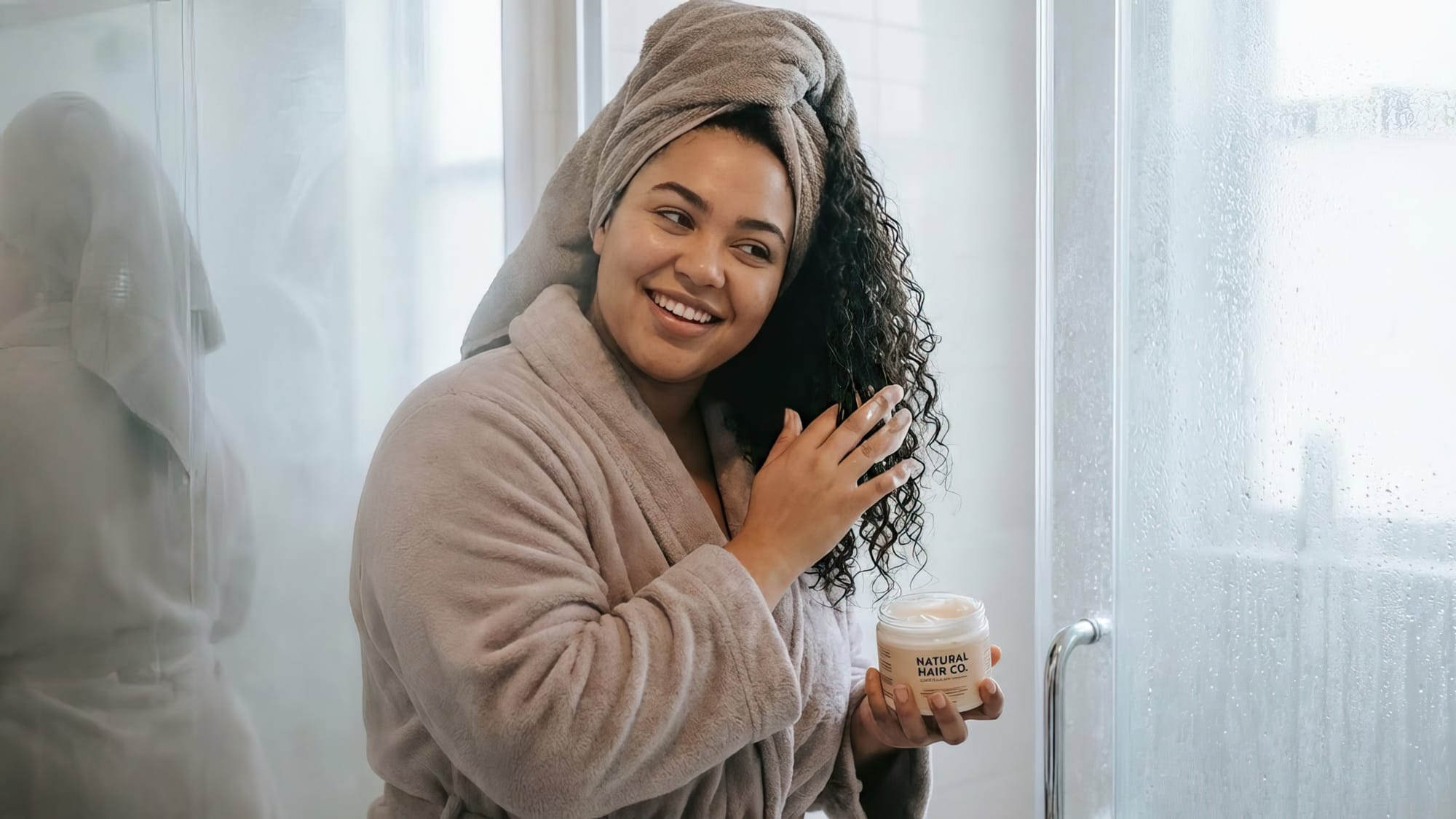 A pretty plus-size woman in her late 20’s with beautiful, healthy-looking hair (wavy or curly texture) in the shower or bathroom, applying natural hair product.