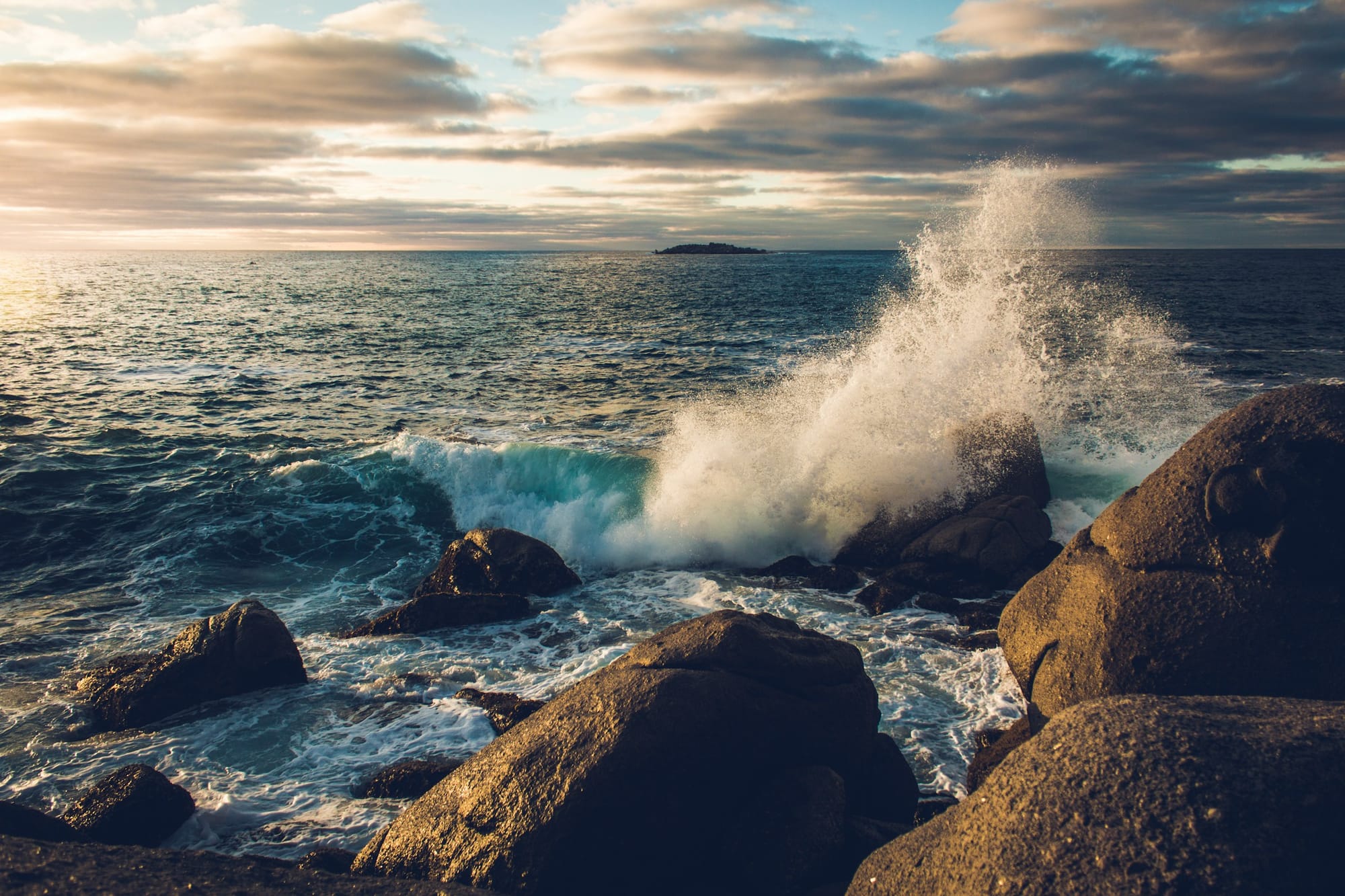A wave breaking against granite boulders at golden hour, its spray lit by the low sun.