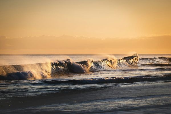 Waves breaking along a beach at golden hour, with backlit spray under a warm orange sky.
