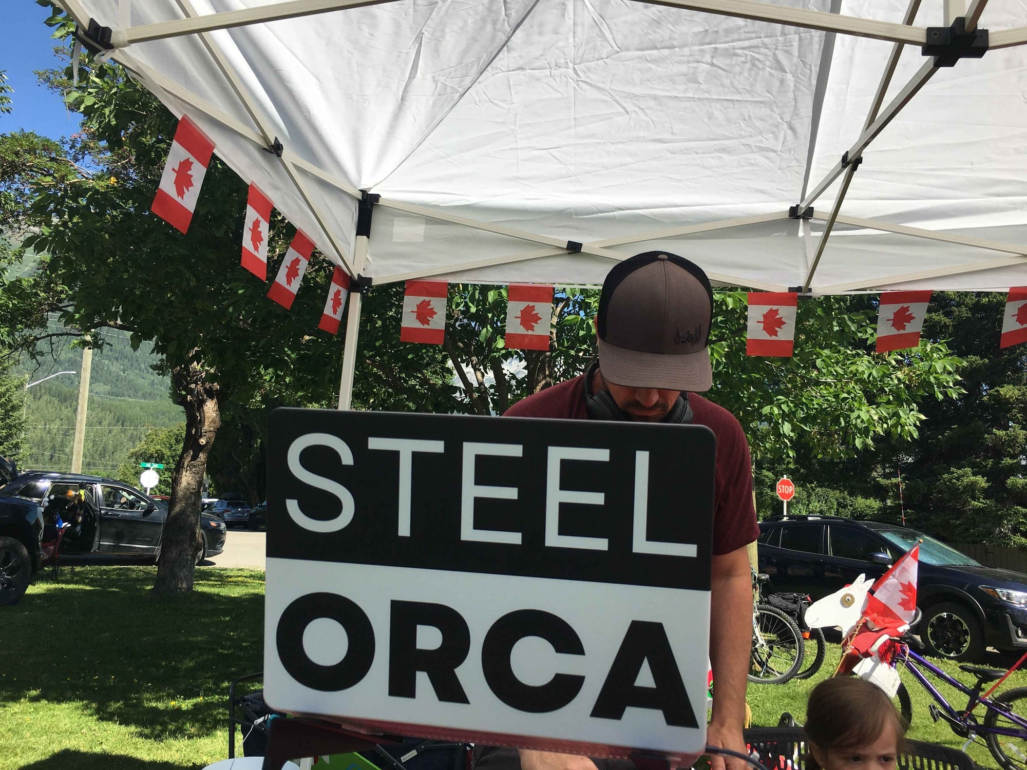 A person wearing a baseball cap looks down behind a black and white sign that reads 'STEEL ORCA'. The setting appears to be an outdoor event, with a white tent overhead decorated with Canadian flags, and cars and trees visible in the background.