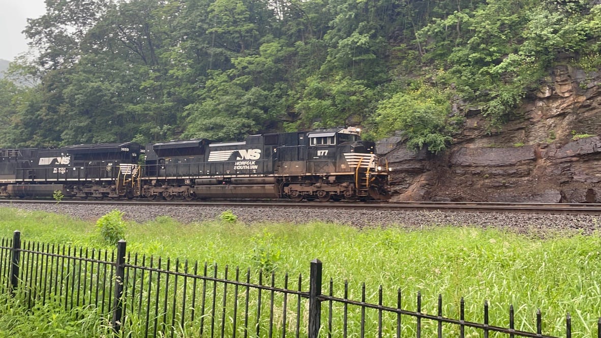 A black and white locomotive with "NS" and the words "Norfolk Southern" beside a horse logo. The train is rounding a curve infront of a cut out hill. There's grass and a black iron fence in the foreground