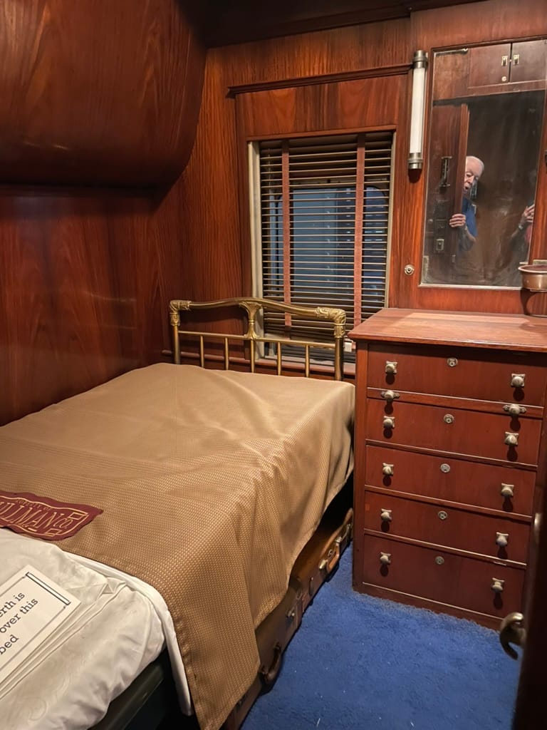 A bed with a golden bedspread and head / endboards made of brass. The walls have dark wood paneling and there's a wooden dresser with a mirror in the background. The carpet is dark blue