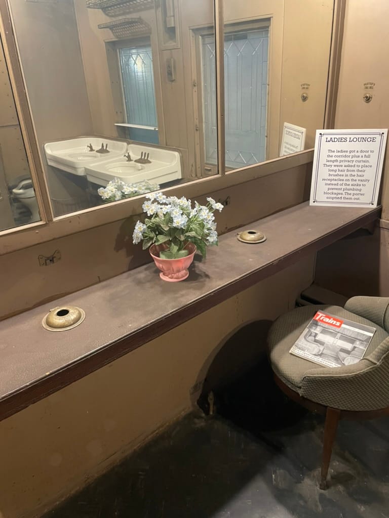 A brown vanity with a mirror. There is a vase with white flowers in the middle, and a chair pulled up to it. The chair has an issue of "Trains" magazine placed on it.