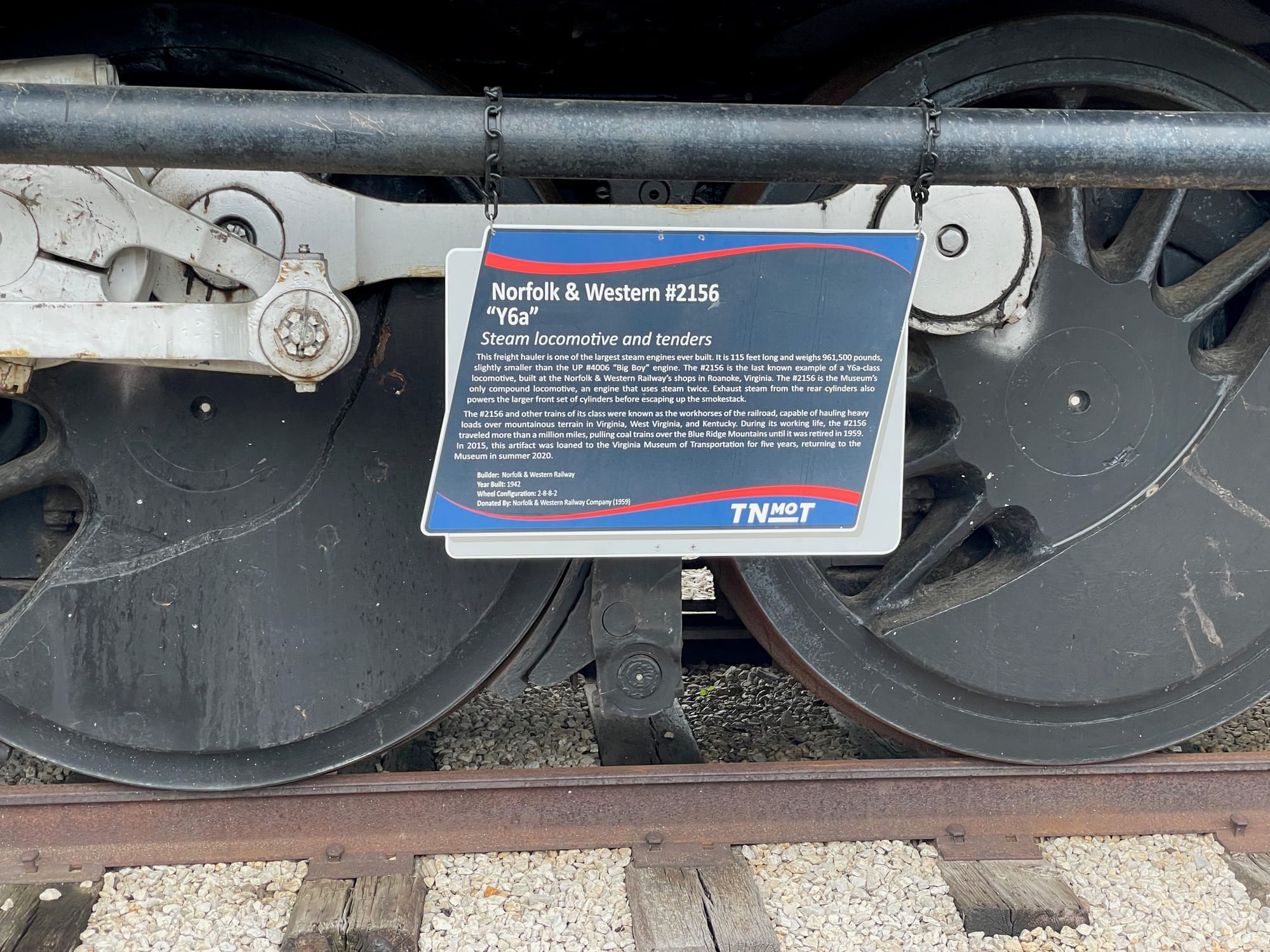 A sign on the wheels of a steam locomotive that reads: Norfolk & Western #2156 "Y6a" Steam locomotive and tenders This freight hauler is one of the largest steam engines ever built. It is 115 feet long and weighs 961,500 pounds, slightly smaller than the UP #4006 "Big Boy" engine. The #2156 is the last known example of a Ya-class locomotive, built at the Norfolk & Western Railway's shops in Roanoke, Virginia. The #2156 is the Museum's only compound locomotive, an engine that uses steam twice. Exhaust steam from the rear cylinders also powers the larger front set of cylinders before escaping up the smokestack. The #2156 and other trains of its class were known as the workhorses of the railroad, capable of hauling heavy loads over mountainous terrain in Virginia, West Virginia, and Kentucky. During its working life, the #2156 traveled more than a million miles, pulling coal trains over the Blue Ridge Mountains until it was retired in 1959. In 2015, this artifact was loaned to the Virginia Museum of Transportation for five years, returning to the Museum in summer 2020. Builder: Norfolk & Western Railway Year Built: 1942 Wheel Configuration: 2-8-8-2 Donated By: Norfolk & Western Railway Company (1959)