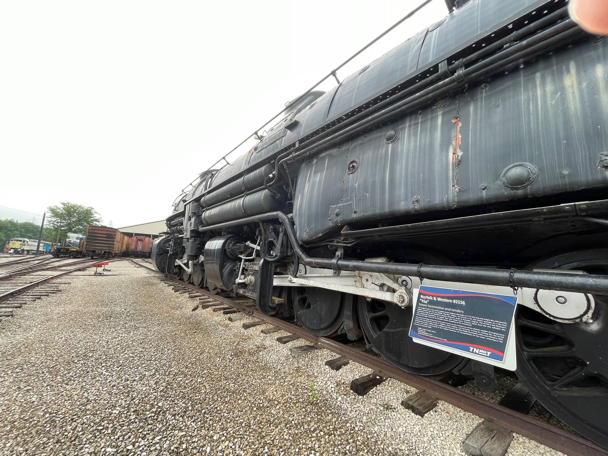 A steam locomotive looking foward from the cab end toward the front. It's a dull rusted black color with silver rods. 3 driving wheels with the silver rods attached in the foreground with the lead drivers forward behind the large piston boxes. There are lots of pipes and handrails running the lengh of the boiler cylander 