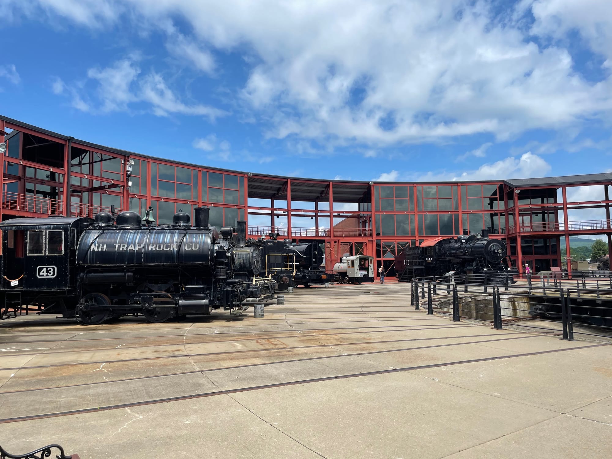 A round concrete area with railroad tracks. On the left the pit of the turntable is visible. There are are several steam locomotives and a round house building with metal frames painted red and glass facades 