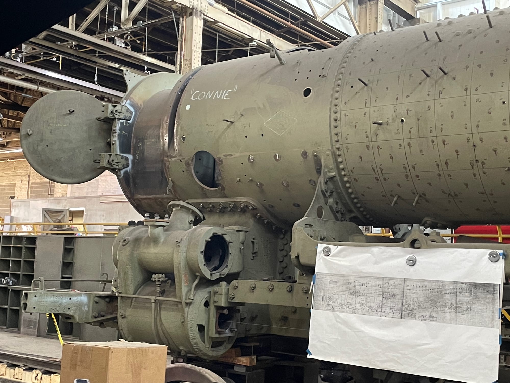 A steam locomotive under construction. The boiler has the jackets removed, exposing the gray/green skin underneath. The forward access hatch is open. The piston shapes are on the locomotive but are incomplete. Bolts and other things are protruding from the surfaces. The wheels are removed.  In the foreground attached to the frame is a schematic the workers are using to put the machine back together.