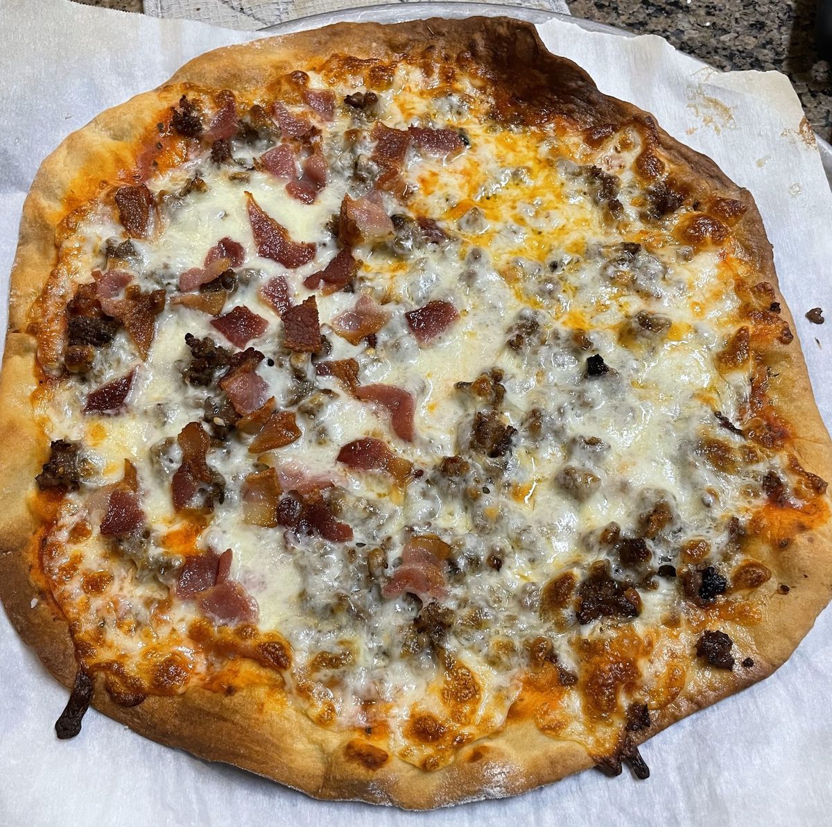 A home made pizza that's not perfectly round. It has browned sausage and bacon on a field of mozzarella cheese. 