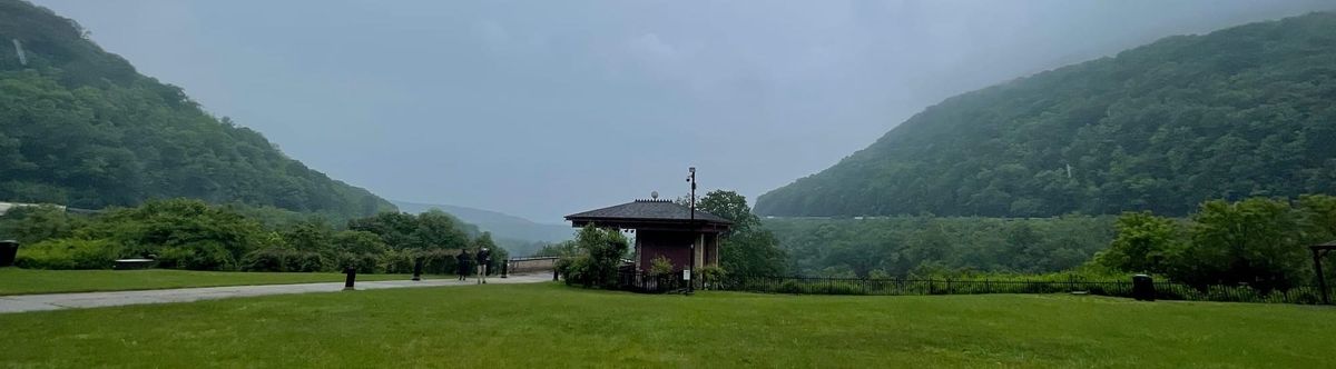 a scene looking down a mountain side. There's a clear cut area with a small building at the center. A train is visible on both mountain sides as it rounds the giant cur