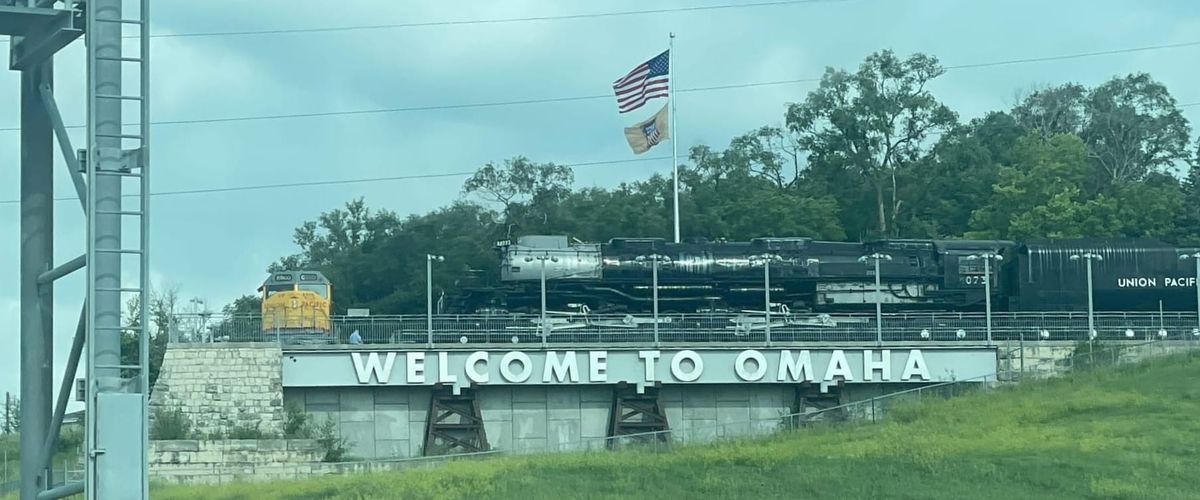 A black and silver steam locomotive facing left to right, on a bluff above a sign that says "Welcome to Omaha". Further left is a yellow diesel electric locomotive facing the camera