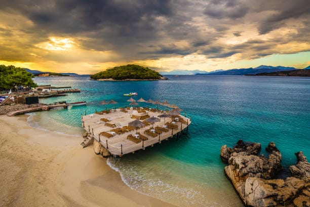 Ksamil beach in Albania with a wooden sunbed deck over turquoise water, sandy shore, and island views under a dramatic sunset sky.
