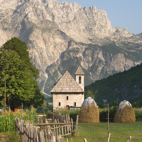 Stone church and haystacks in Theth National Park, surrounded by Albania’s Albanian Alps mountain range.