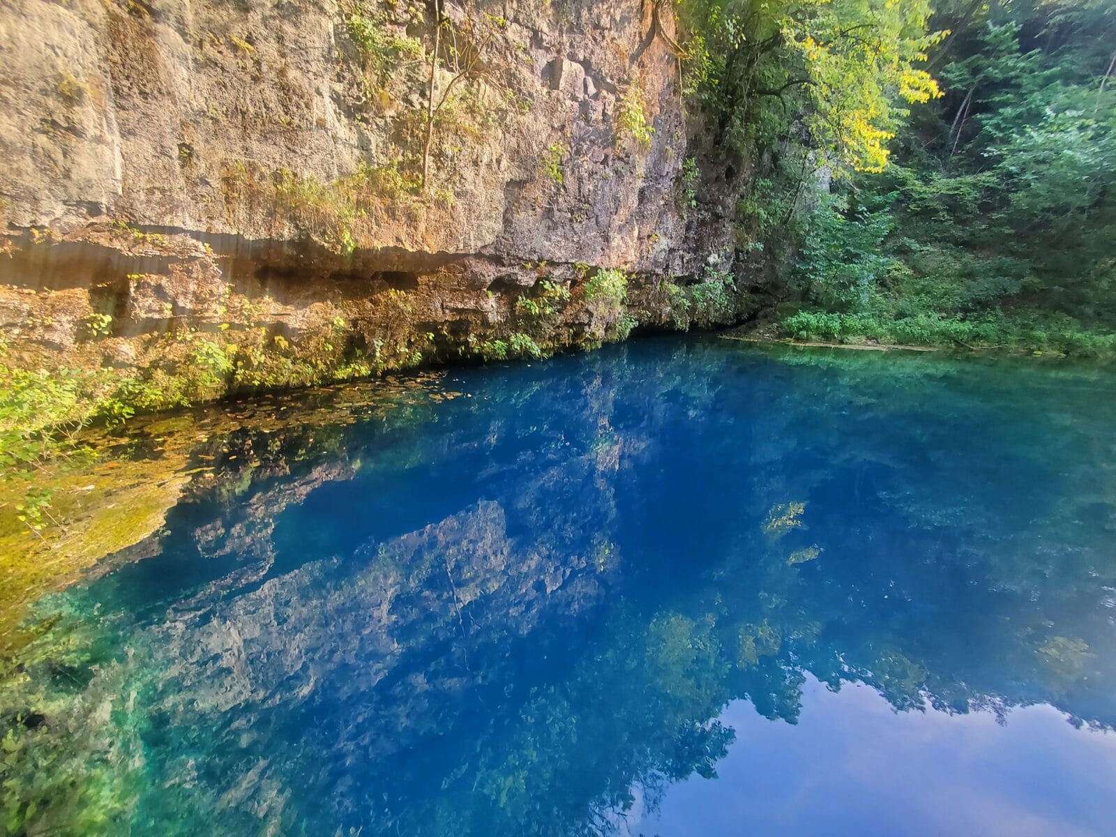 Bright blue deep spring with a large rock cliff on he side of it.