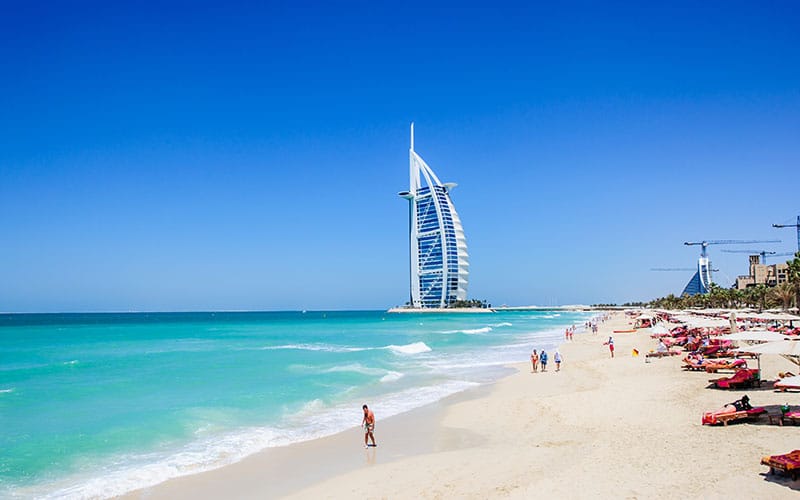 A view of the sailboat 7 star hotel in Dubai from a beautiful beach and clear blue waters with people handing out playing in the sand. 