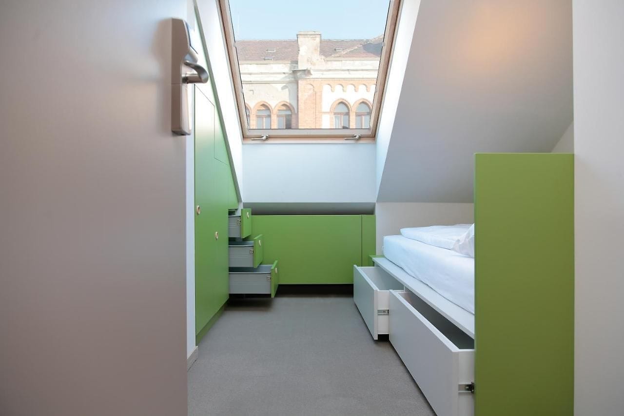 A room with a single twin bed with green accents in  the room and a window offering views of the street in Budapest. 