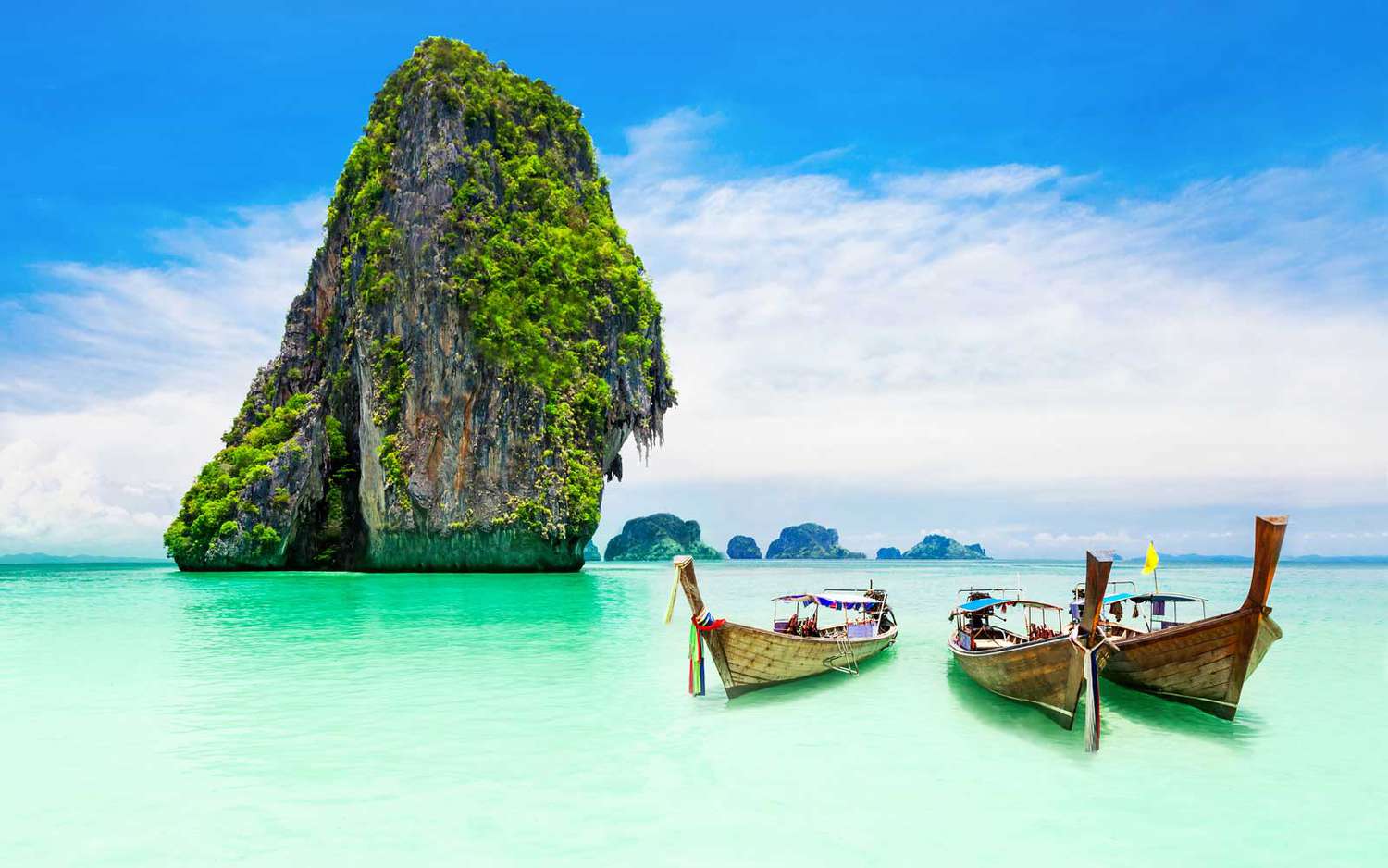 View of boats sitting in the water with a large rock popping up out of the sea in Phuket, Thailand. 