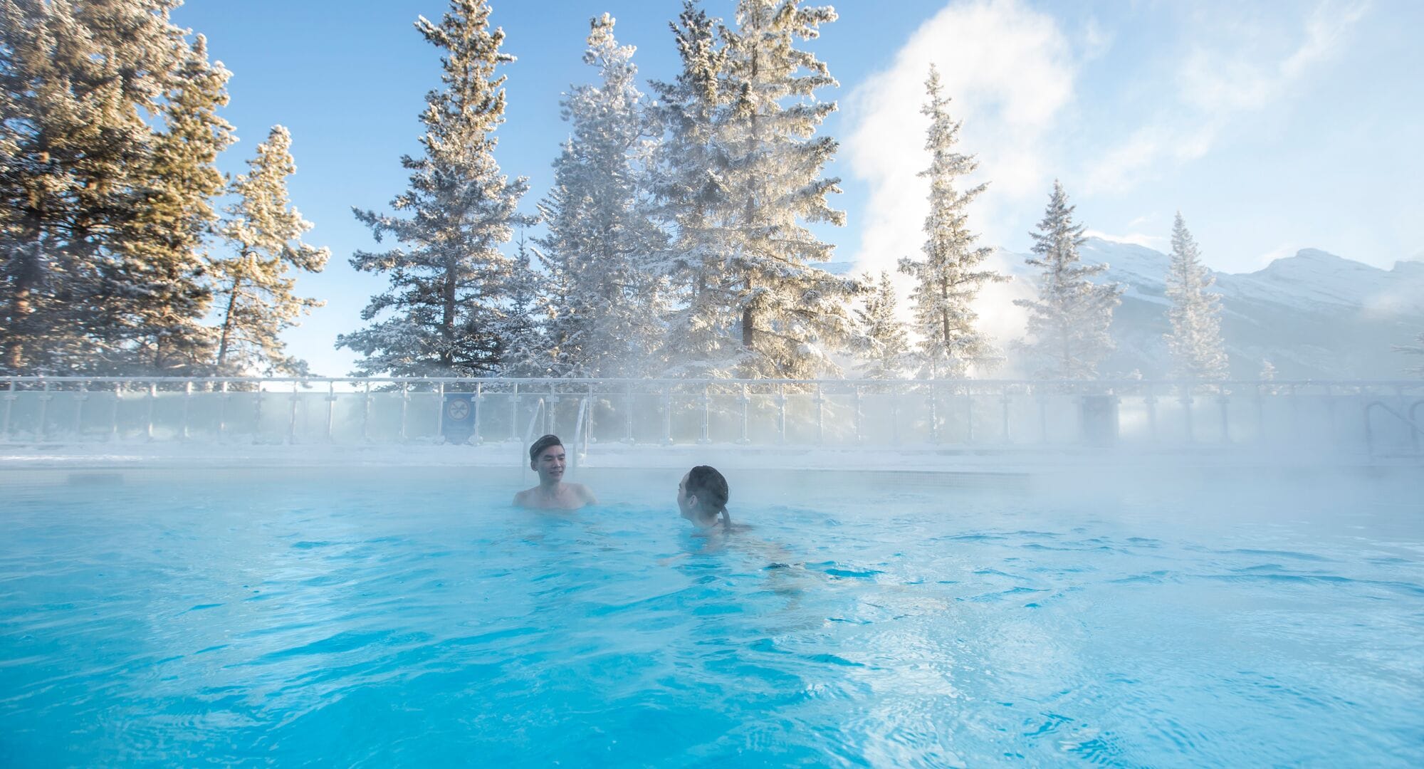 Two people sitting in a hot spring with steam coming off of it and surrounded by snow covered pine trees with clear blue skies.