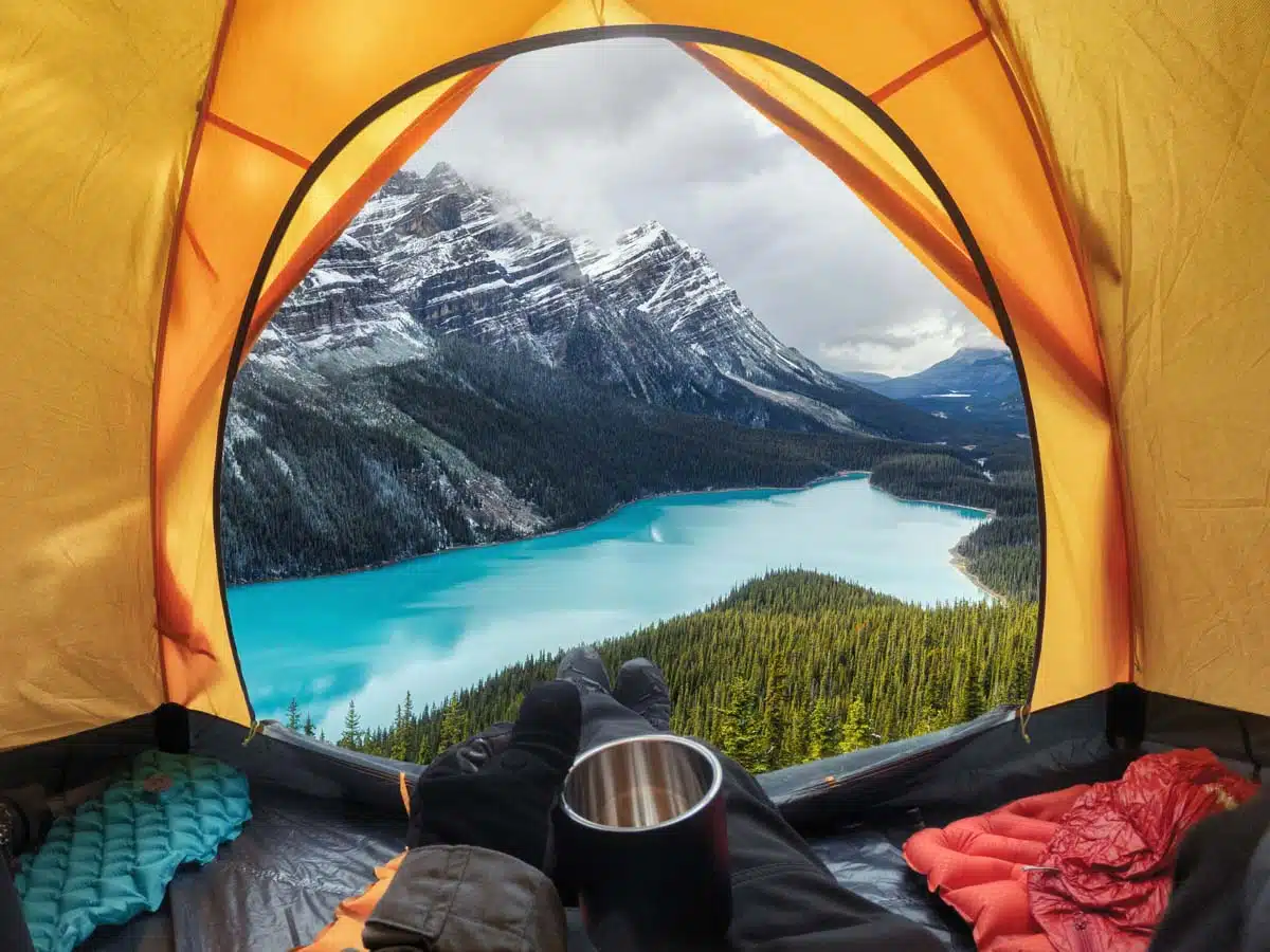 A person inside of a yellow tent with a view looking outside the tents door showing a beautiful lake and mountain below their campsite.