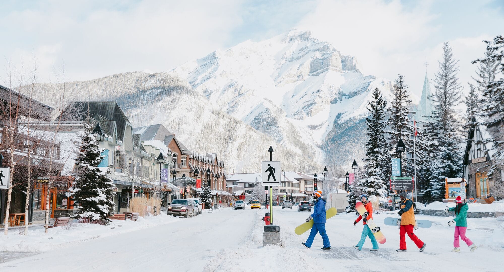 People walking with snowboards across a snow covered street inside Banff National Park.