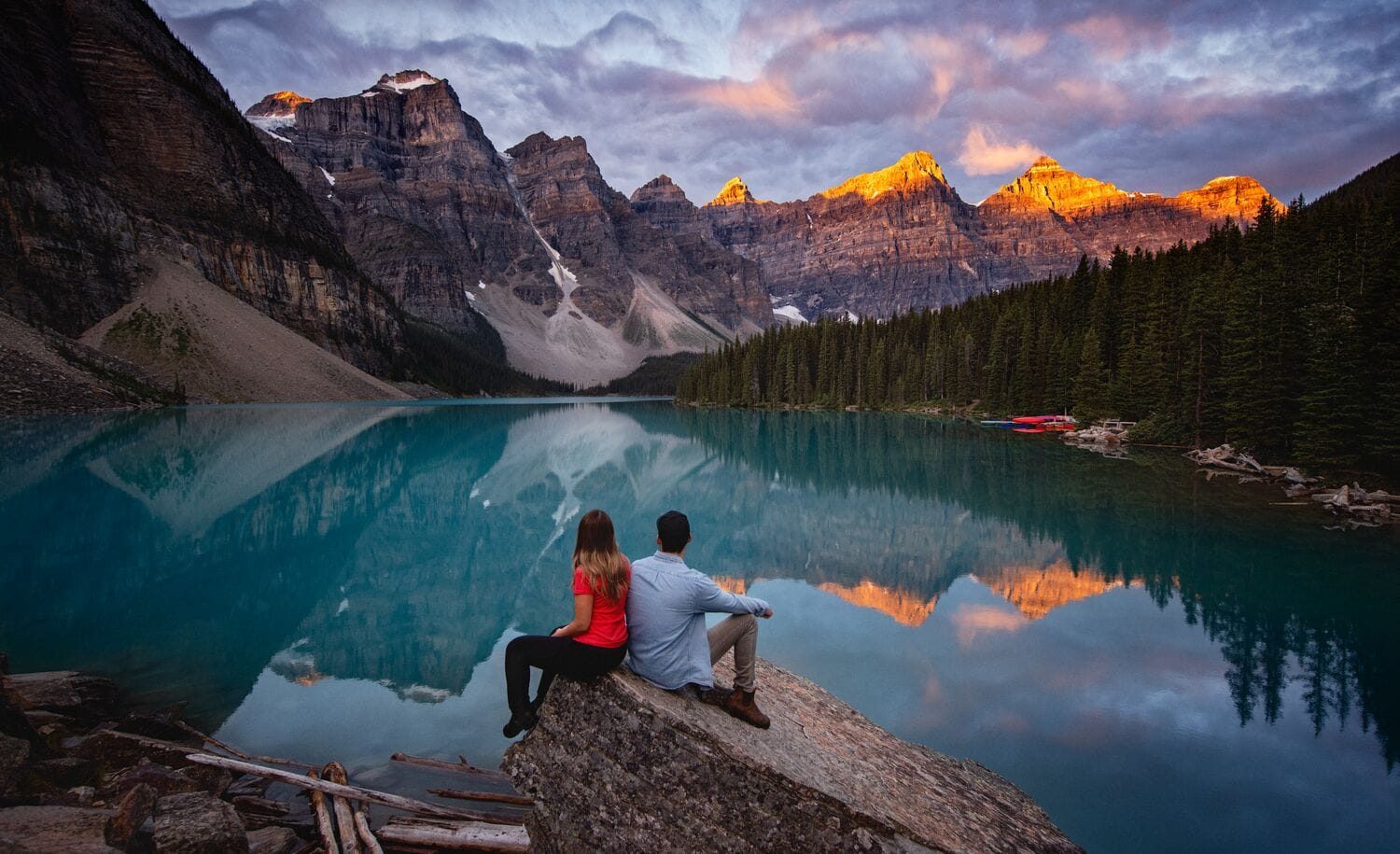 Two people sitting on a rock over looking a clear blue lake that is surrounded by pine trees and mountains.
