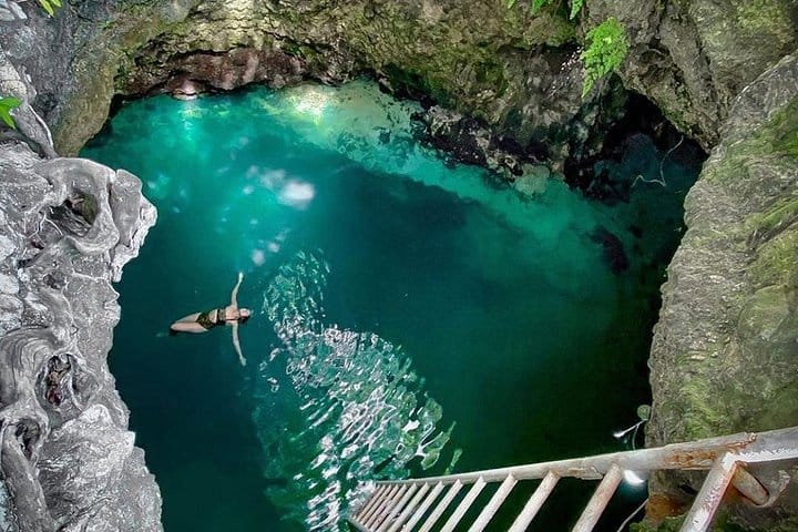 A person diving into a beautiful clear pool of water with cliffs on all sides forming a circle and a ladder down in the water to climb back up at the Blue hole Mineral Spring in Negril.