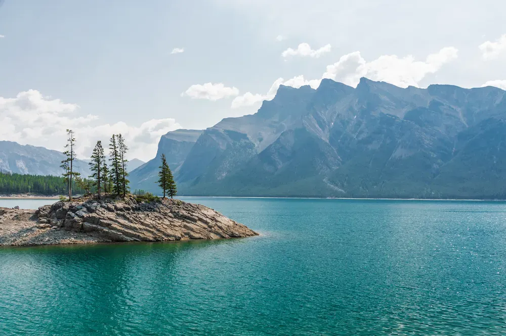 A pretty lake with a rock island sitting in the middle with pine trees and in the distance the lake ends at the base of a large mountain. 