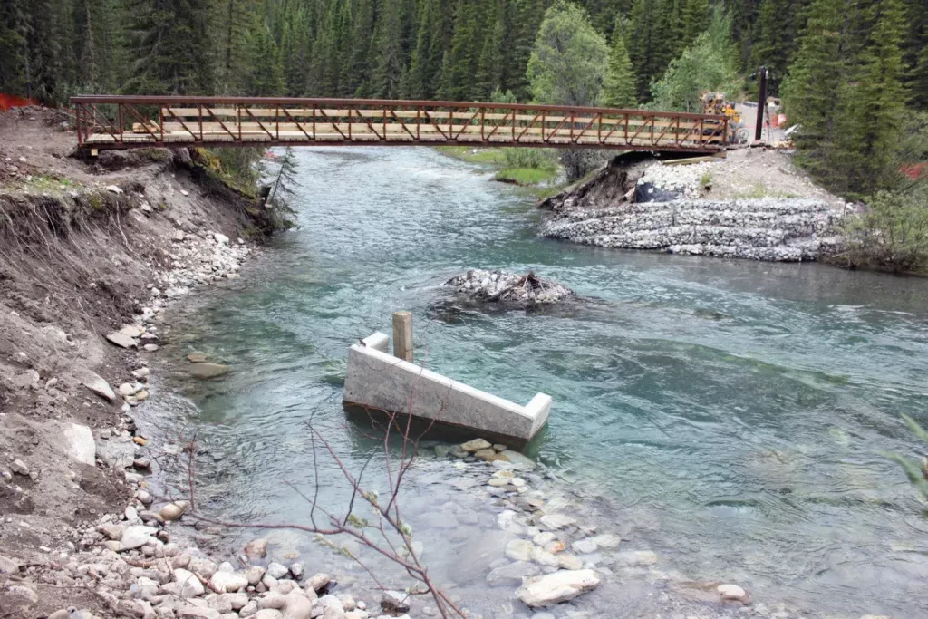 A Pedestrian bridge crossing over a shallow narrow river with rocky banks and surrounded by pine trees.