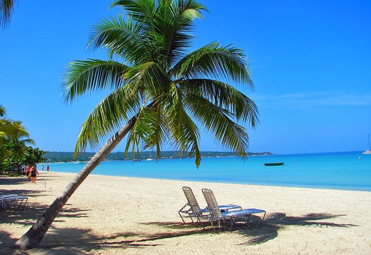 A palm tree hanging over beach chairs and a view of clear Caribbean waters at Negril's Half Moon Beach.