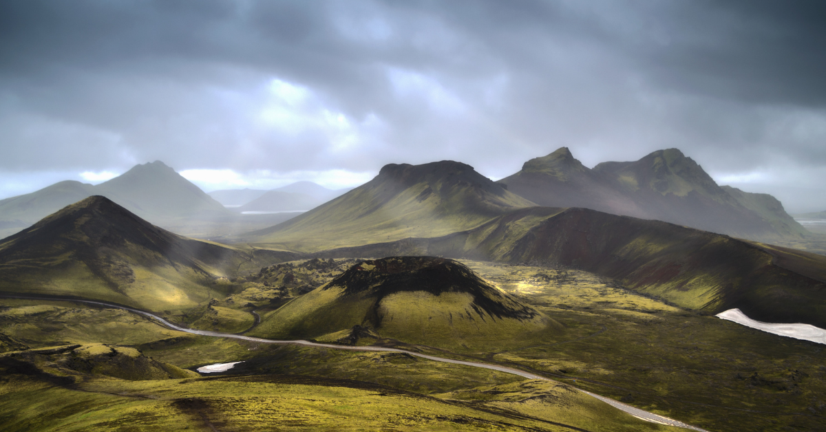 Many rugged mountains spread out across Iceland's green plains and a river running in front of then.