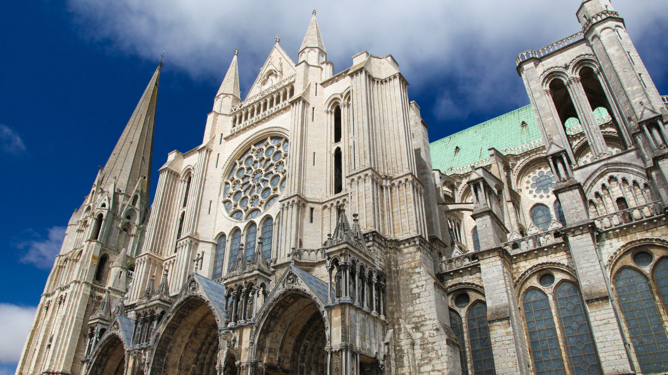 Gothic façade of Chartres Cathedral in France, featuring intricate stone carvings, tall spires, and a large rose window under a bright blue sky with scattered clouds.