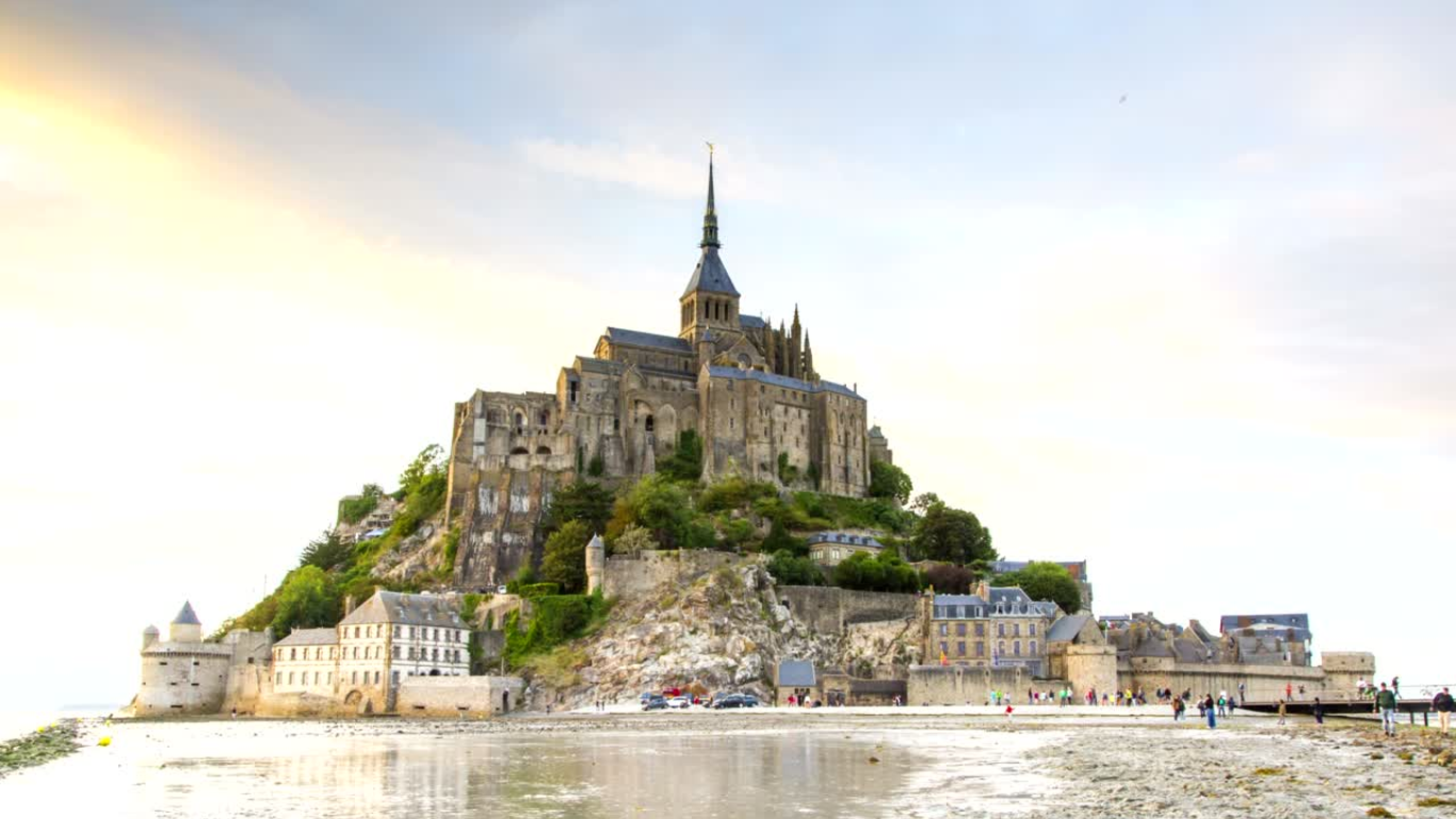 Mont-Saint-Michel in Normandy, France, at low tide, showing the iconic medieval abbey perched atop the rocky island with surrounding stone buildings and visitors walking along the exposed bay.