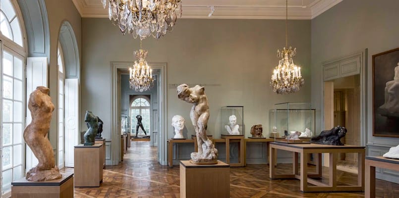 A white room with chandeliers and statues places around the room and artwork on the walls. 