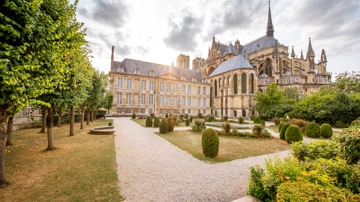 Rear view of the Notre-Dame de Reims Cathedral in France, with Gothic architecture and a landscaped garden featuring trimmed trees, pathways, and manicured bushes under a partly cloudy sky.