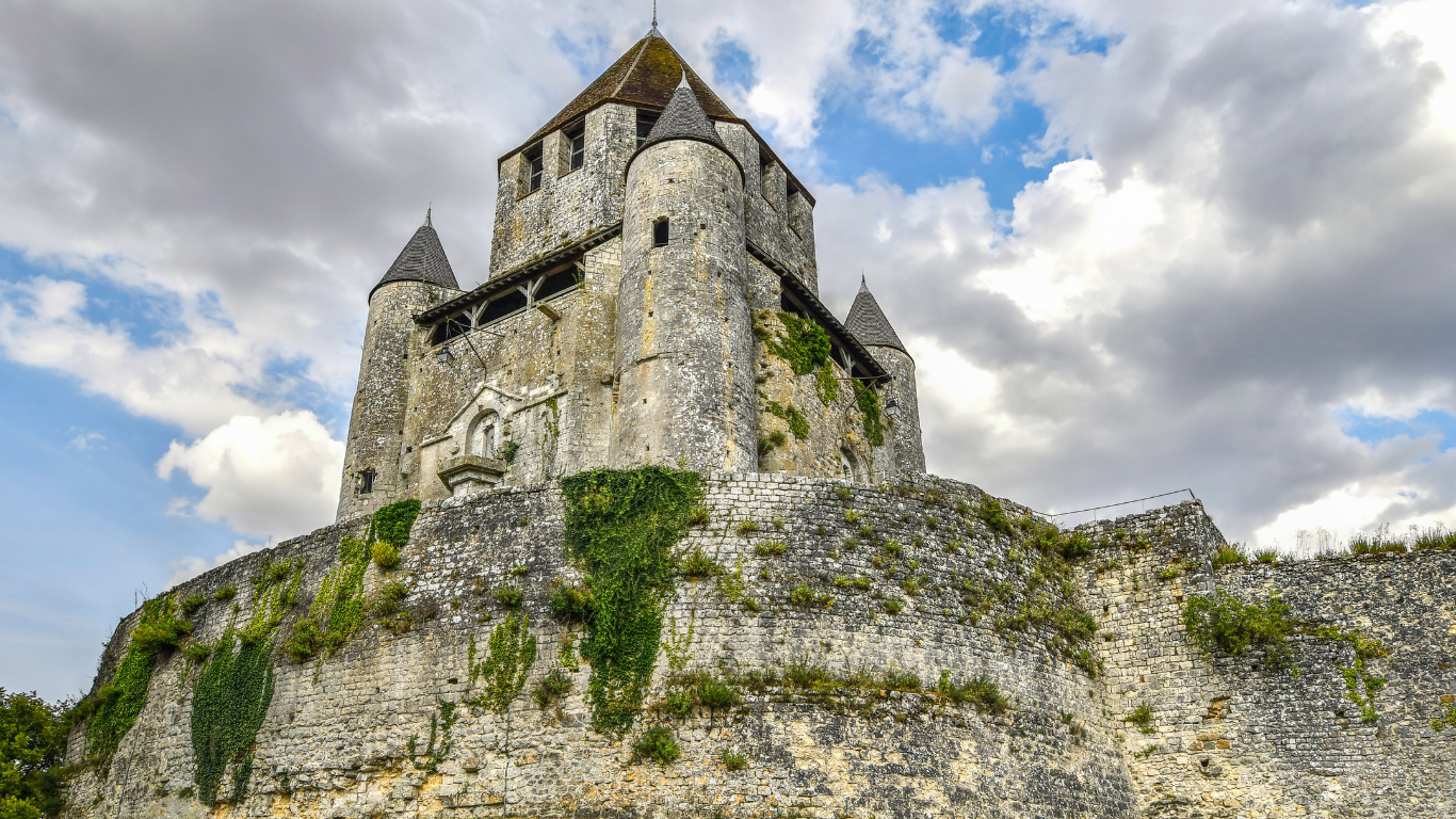 Medieval stone castle in Provins, France, with round towers and ivy-covered walls, perched on a hilltop under a dramatic cloudy sky.