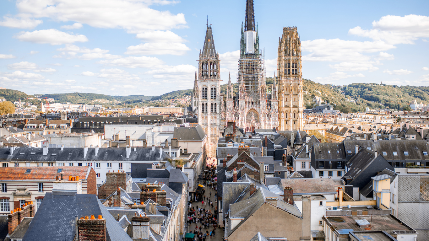 Aerial view of Rouen Cathedral in Normandy, France, with its Gothic spires rising above the rooftops of the city center, surrounded by historic buildings and rolling hills under a partly cloudy sky.