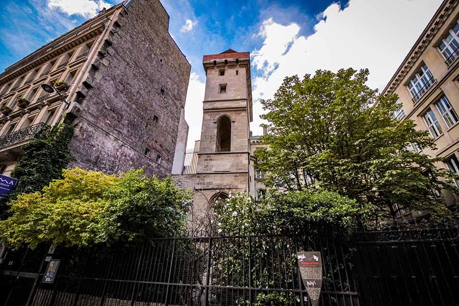 Tour Jean-sans-Peur, a medieval tower in Paris, France, surrounded by greenery and historic buildings, with a dramatic sky overhead.