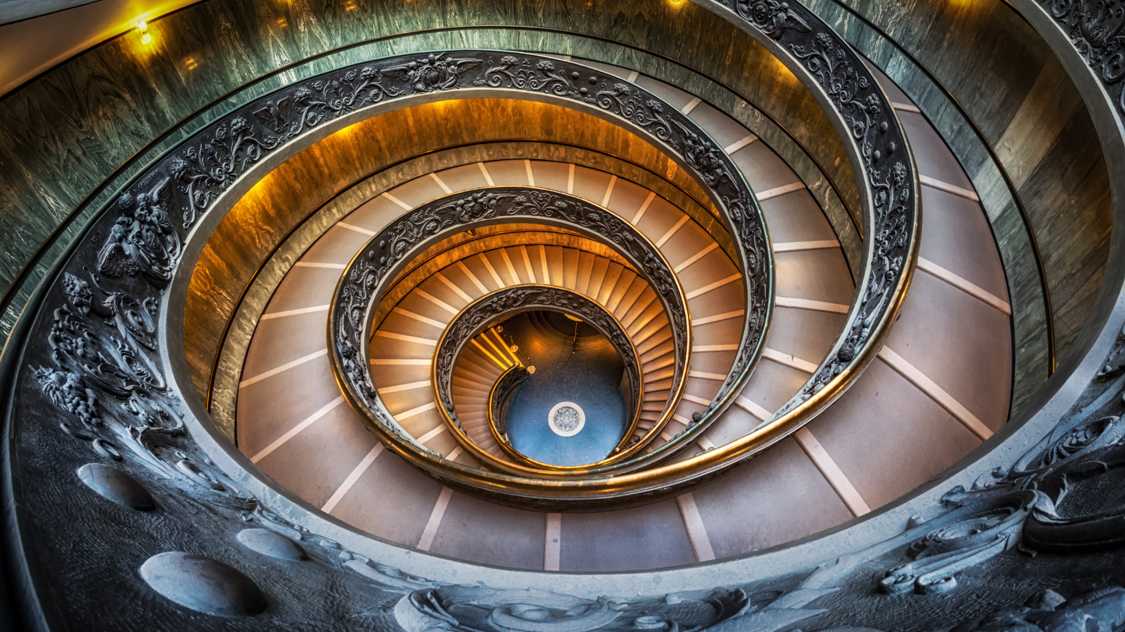 A top-down view of the iconic Bramante Staircase in the Vatican Museums, Vatican City. The double-helix spiral staircase features intricate bronze railings adorned with decorative reliefs and floral motifs. 
