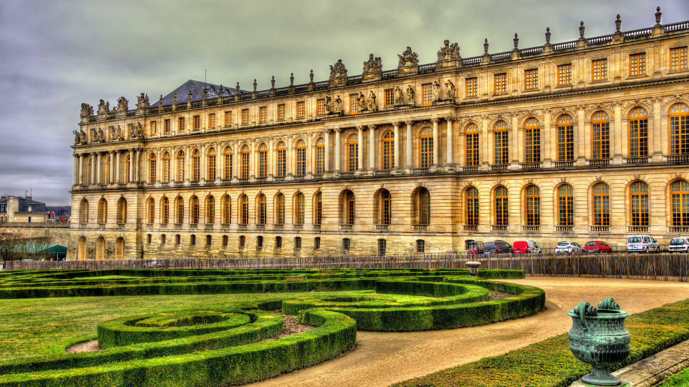 Elegant façade of the Palace of Versailles in France, with ornate architecture and meticulously trimmed geometric hedges in the foreground of the formal gardens.