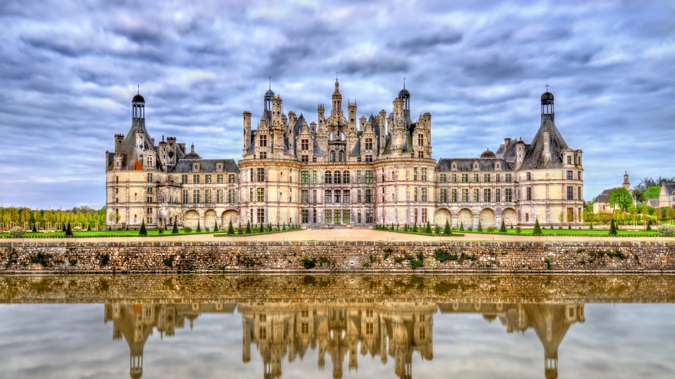 The Château de Chambord in France, featuring intricate Renaissance architecture with towers and chimneys, reflected in a still moat under a dramatic cloudy sky.
