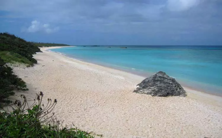 A beach with a large rock near the waterfront and the skies are dark and looks like rain is coming. 