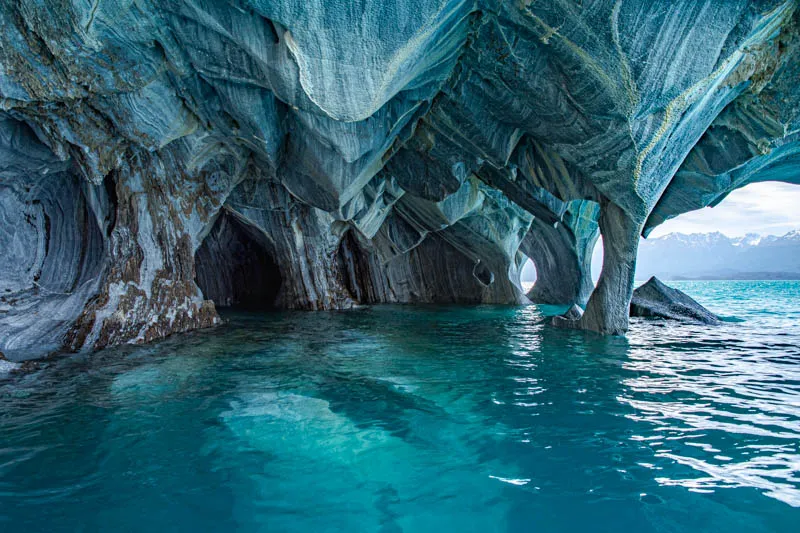 Expansive view of the Marble Caves in Patagonia, Chile, with swirling blue and gray marble formations rising above vibrant turquoise water and distant snow-capped mountains on the horizon.
