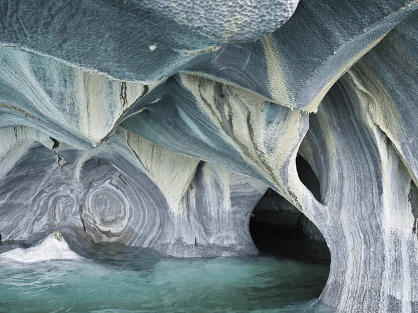 Interior view of the Marble Caves in Patagonia, Chile, featuring smooth, swirling gray and white marble walls above light green-blue water, with dark cave openings in the background.